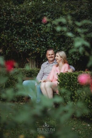 Engagement Photography: a couple sit on a park bench against a green hedge