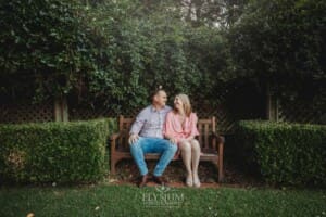 Engagement Photography: a couple sit on a park bench against a green hedge