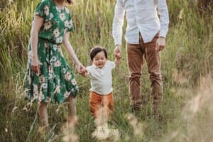 A family hold their baby's hands as he walks with them