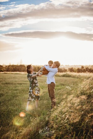 A family stand on a grassy hill at sunset cuddling their baby boy
