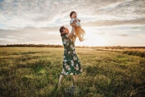 A mother lifts her baby boy into the air on a grassy hill