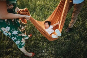 A baby boy sits in a blanket hammock as his family swing him
