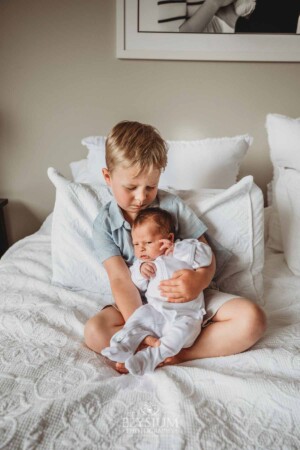 A boy cuddles his newborn sister as they sit on a bed