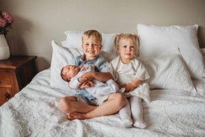 Siblings hold their baby sister as they sit on a white bed
