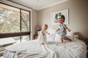 Siblings jump on a white bed in their parents room
