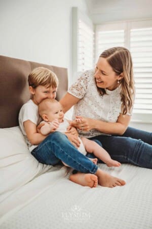 Newborn Photography: a little boy sits on a bed and cuddles his baby sister in his lap