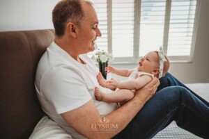 Newborn Photography: a father sits on a bed with his baby girl in his lap