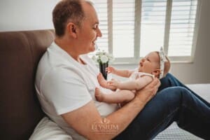 Newborn Photography: a father sits on a bed with his baby girl in his lap