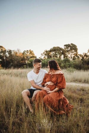 Outdoor Session - a man sits with his pregnant wife in long grass at sunset