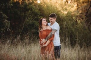 A man hugs his pregnant wife in a long linen maxi dress standing in a grassy field