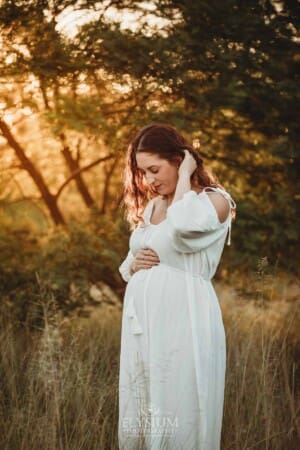 Outdoor Session - a pregnant woman sweeps hair behind her ear in a white maternity dress
