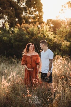 A man holds hands with his pregnant wife standing in long grass at sunset