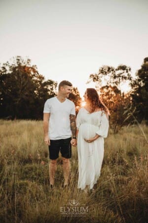 Outdoor Session - a man holds his pregnant wife's hand as they stand in long grass