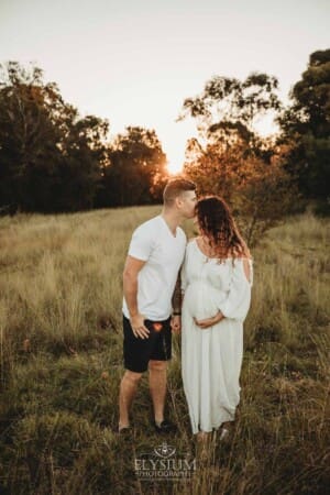 Outdoor Session - A man kisses his pregnant wife who's wearing a white maternity gown