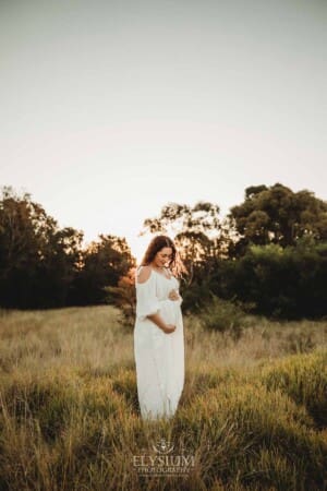 Outdoor Session - A pregnant woman stands in long grass in a white maternity gown