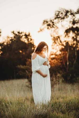 Outdoor Session - A pregnant woman stands in long grass in a white maternity gown