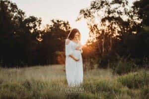 Outdoor Session - A pregnant woman stands in long grass in a white maternity gown