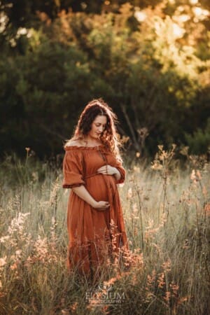 A pregnant woman wearing a long rust maternity dress holds her pregnant belly at sunset