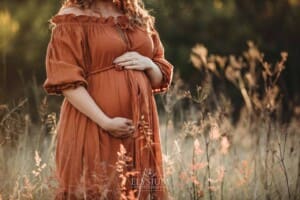 Outdoor Session - a woman wearing a long rust maternity dress holds her pregnant belly
