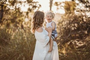 A pregnant woman sits her child on her belly while she wears a long white linen dress