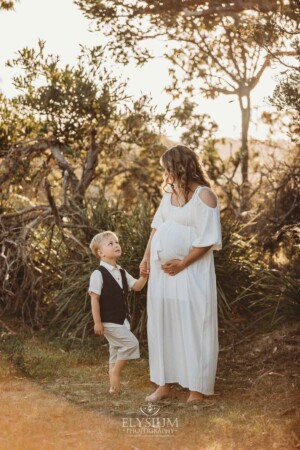 A pregnant woman in a white maternity gown stands holding her childs hand at sunset