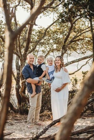 A pregnant woman in a white maxi dress stands on a beach with her family surrounded by trees