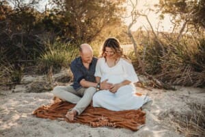 A pregnant mother sits with her partner on an amber blanket in a long white maternity gown