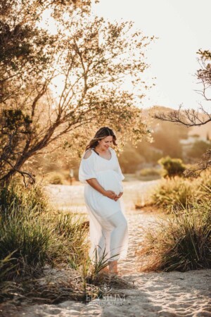 A pregnant woman in a white linen dress stands on a sandy beach at sunset