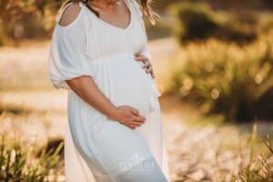 A pregnant woman in a white linen dress stands on a sandy beach at sunset