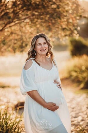 A pregnant woman in a white linen dress stands on a sandy beach at sunset