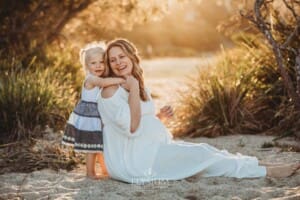 A pregnant woman sits on the sand with her child cuddling her at sunset