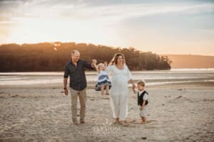 A pregnant woman in a long white dress walks across the sand with her family at sunset