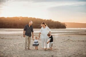 A pregnant woman in a long white dress walks across the sand with her family at sunset