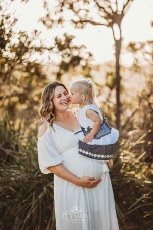 A pregnant woman in a white maxi dress holds her child as she kisses her mother on the cheek