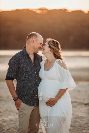 A pregnant mother in a white dress rubs noses with her partner as the sun sets behind them on a beach