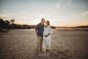 A pregnant woman stands on a beach with her partner at sunset