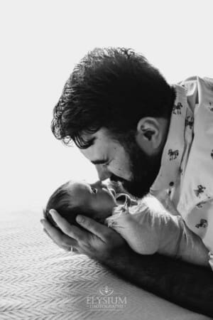 A father holds one of his newborn twins in his hands and lays on a white blanket