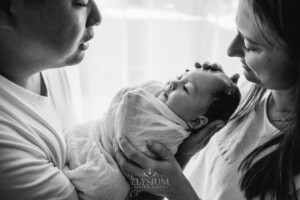 Parents hold their newborn baby boy wrapped in a soft white blanket and look adoringly at him