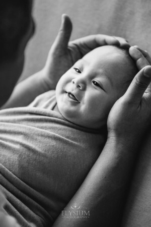 A father holds his smiley baby boy in his hands as he lays on a blanket, black and white photograph