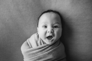 A smiley happy newborn baby smiles as he lays in a wrap on a matching blanket
