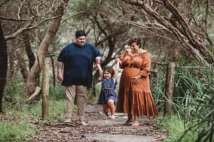 Parents walk a beach path holding their daughter's hand with the mother in an amber linen maternity gown