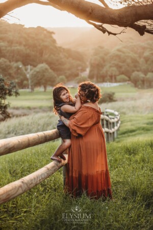 A mother cuddles her baby girl at sunset as she sits on a wooden fence in a grassy field
