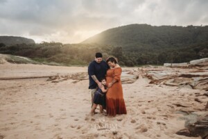 Parents cuddle their baby girl as they stand on a beach with the sun setting behind them