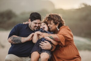 Parents tickle their little girl as she sits on their laps giggling with the sun setting behind them