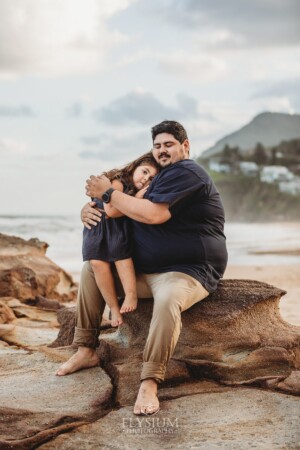 A little girl sits in her dads lap and cuddles him as they sit on beach rocks at sunset