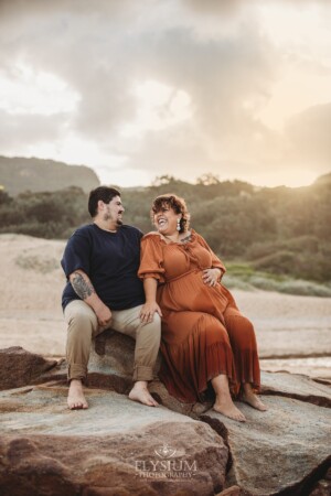 A pregnant woman cuddles up to her partner as they sit on beach rocks at sunset