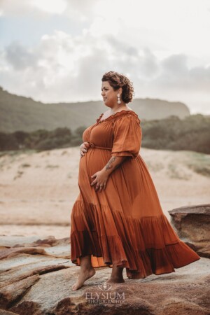 A pregnant woman in an amber maternity gown stands on beach rocks at sunset