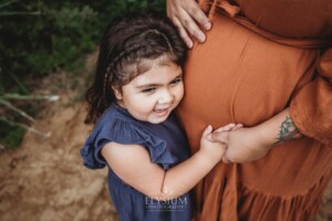 A little kid in a navy linen dress cuddles her mums pregnant belly in an amber maternity gown and giggles