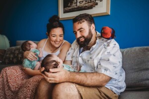 Parents sit holding their newborn baby twins with their parrot on dads shoulder