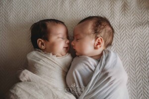 Newborn twins lay on a textured white blanket wrapped with their little noses pressed together